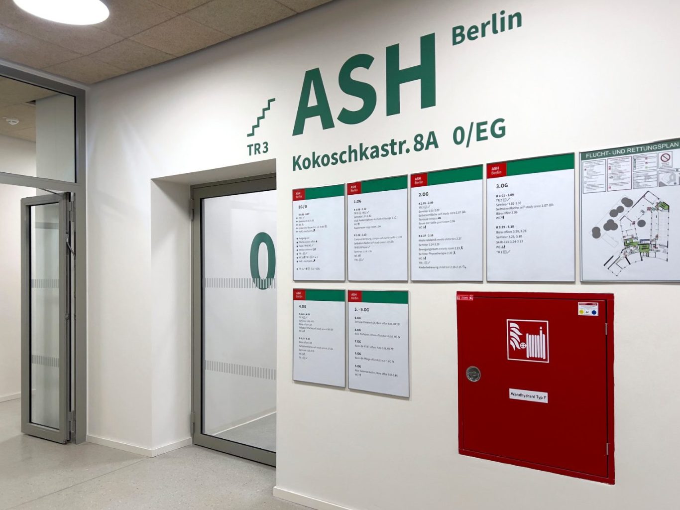 Gestaltung Etagenübersicht Foyer Wegeleitsystem Alice-Salomon Hochschule Berlin Wand mit Informationen der ASH Berlin, einschließlich Notfallhinweisen und Plänen.
