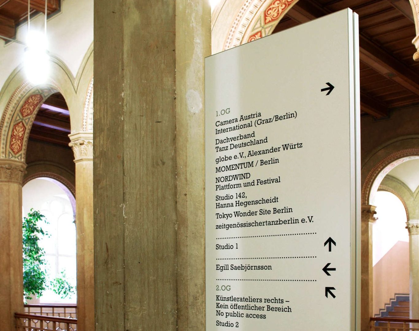 Gestaltung Richtungsbeschriftung auf Stele Orientierungssystem Kunstzentrum Bethanien Berlin Kreuzberg Helle Informationsstele in einem historischen Gebäude mit Wegweisern.